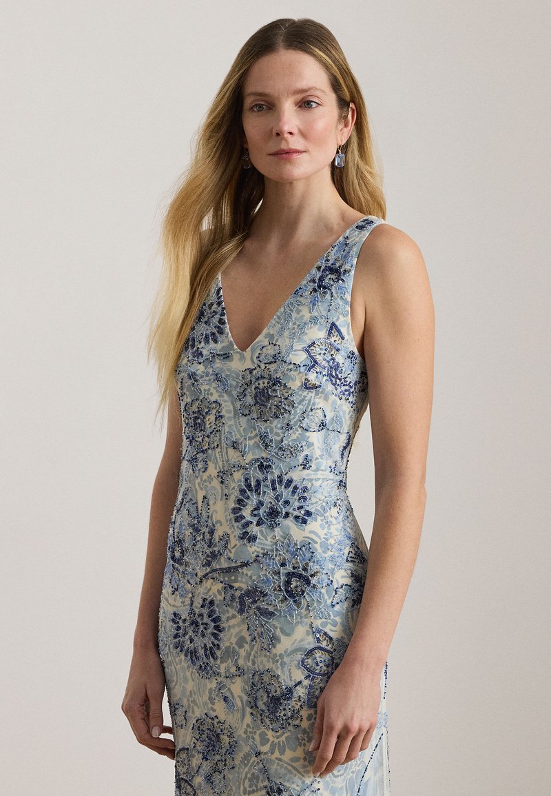 Γυναίκα με μακριά ξανθά μαλλιά που φοράει αμάνικο φόρεμα με λαιμόκοψη V και μπλε κεντήματα με λουλούδια, μπροστά σε ένα μονόχρωμο φόντο.