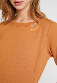 Vêtement orange brûlé avec un col rond, présentant un détail de couture subtil et quatre boutons décoratifs près de l'épaule.