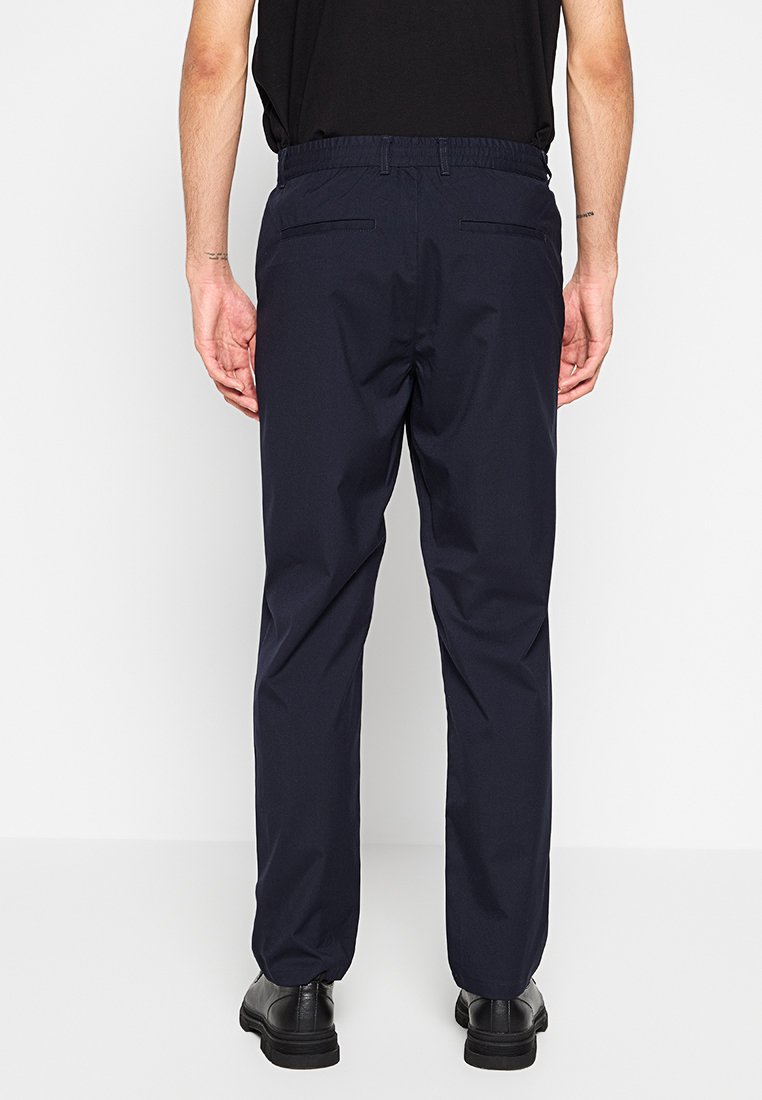 Marineblaue Hose mit elastischem Bund und lockerer Passform, zwei Gesäßtaschen, getragen mit einem schwarzen Kurzarmhemd und schwarzen Schuhen.