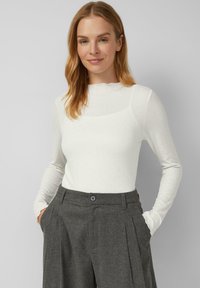 Weißes Langarmoberteil mit einem gewellten Ausschnitt und zartem Texturmuster. Kombiniert mit grauen, hochgeschnittenen Hosen mit Falten und Taschen.