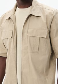 Jachetă bej cu mâneci scurte, cu fermoar frontal, două buzunare pe piept și un tricou subțire de un bej deschis, texturat. Material neted cu un design casual.