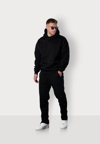 Schwarzer Kapuzenpullover aus weichem Material, ausgestattet mit einer Kängurutasche. Kombiniert mit schwarzen Jogginghosen und weißen Sneakers.