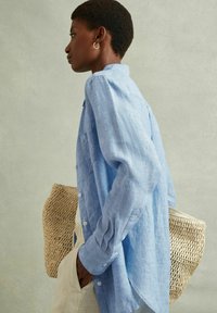 Camicia button-up in lino azzurro chiaro con maniche lunghe, caratterizzata da una tasca frontale, indossata sopra pantaloni beige; il modello tiene una borsa di paglia intrecciata.