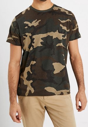 Mężczyzna ubrany w koszulkę z krótkim rękawem w kamuflażowy wzór oraz beżowe spodnie, stojący z rękami swobodnie opuszczonymi wzdłuż ciała.