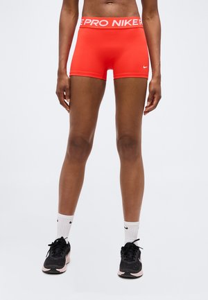 Sportos személy élénk narancssárga Nike Pro rövidnadrágot, fehér Nike zoknit és fekete sportcipőt visel, világosszürke háttéren állva.