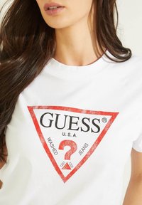 Balta kokvilnas T-kreklu, kurā redzams sarkans trīsstūris logotips ar uzrakstiem "GUESS," "U.S.A." un "WASHED JEANS" melnā krāsā. Papildu dizaina elementi ietver jautājuma zīmi.