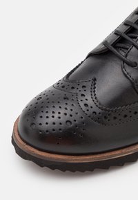 Černá kožená bota s designem brogu, perforované detaily na špičce, kontrastní hnědá podrážka a texturované šněrování.