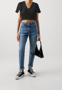 Černý texturovaný crop top s krátkými rukávy, v kombinaci s vysokými světle modrými džíny a černými plátěnými teniskami. Drží malou černou kabelku.