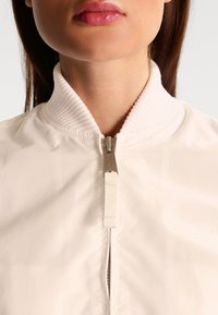 Veste bomber beige clair avec un col côtelé, une fermeture éclair frontale complète et une finition en tissu lisse et brillant. Dispose d'un petit logo sur le tirette de la fermeture éclair.