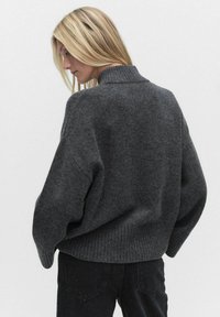 Donkergrijs, oversized gebreide trui met hoge hals en geribbelde zoom. Zachte textuur, verlaagde schouders en gedetailleerde stiksels op de achterkant.