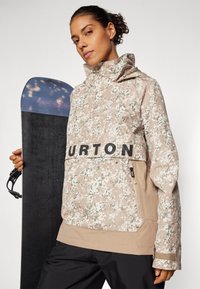 Jasnozłota i zielona kurtka w kamuflażu o lekkiej wadze, z wysokim kołnierzem i czarnym logo "BURTON"; posiada kieszeń na zamek błyskawiczny i elastyczne mankiety.