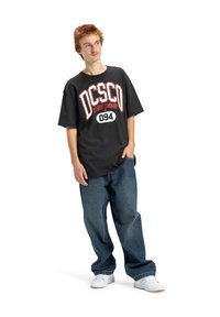 Čierne oversize tričko s bielym a červeným logom "DC Shoe Company" a číslom "094"; skombinované s voľnými modrými džínsami a bielymi teniskami.