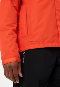 Giacca impermeabile rossa con finitura testurizzata e polsini a velcro. Presenta il marchio "CEPLEX ACTIVE", abbinata a pantaloni neri con logo "VAUDE".