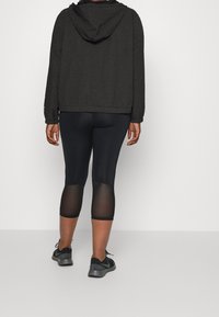 Zwarte, gestructureerde hooded jacket gecombineerd met zwarte capri-legging met doorzichtige panelen, aangevuld met donkere sportschoenen.