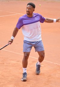 Tennisutøver iført en lila og hvit kortermet skjorte og grå shorts, som holder en racket på en leirk Court. Grå treningssko og hvite sokker.