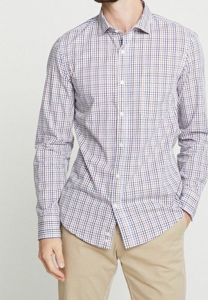 Mężczyzna w dopasowanej, zapinanej na guziki koszuli z długim rękawem w niebiesko-brązową kratę i beżowych spodniach, z ręką w kieszeni.