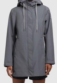 Šedá kapucňová bunda s bílými šňůrkami, předními kapsami a patentními knoflíky, nošená přes černé kraťasy na osobě s tmavou pletí.