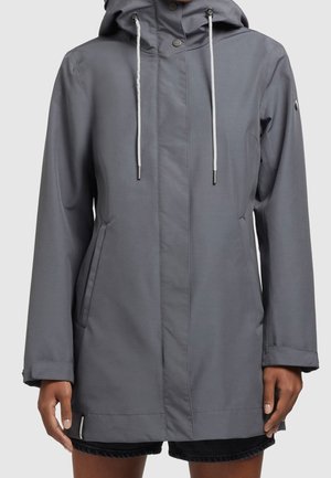 Grå huva jacka med vita dragband, framfickor och tryckknappar som bärs över svarta shorts på en person med mörk hy.