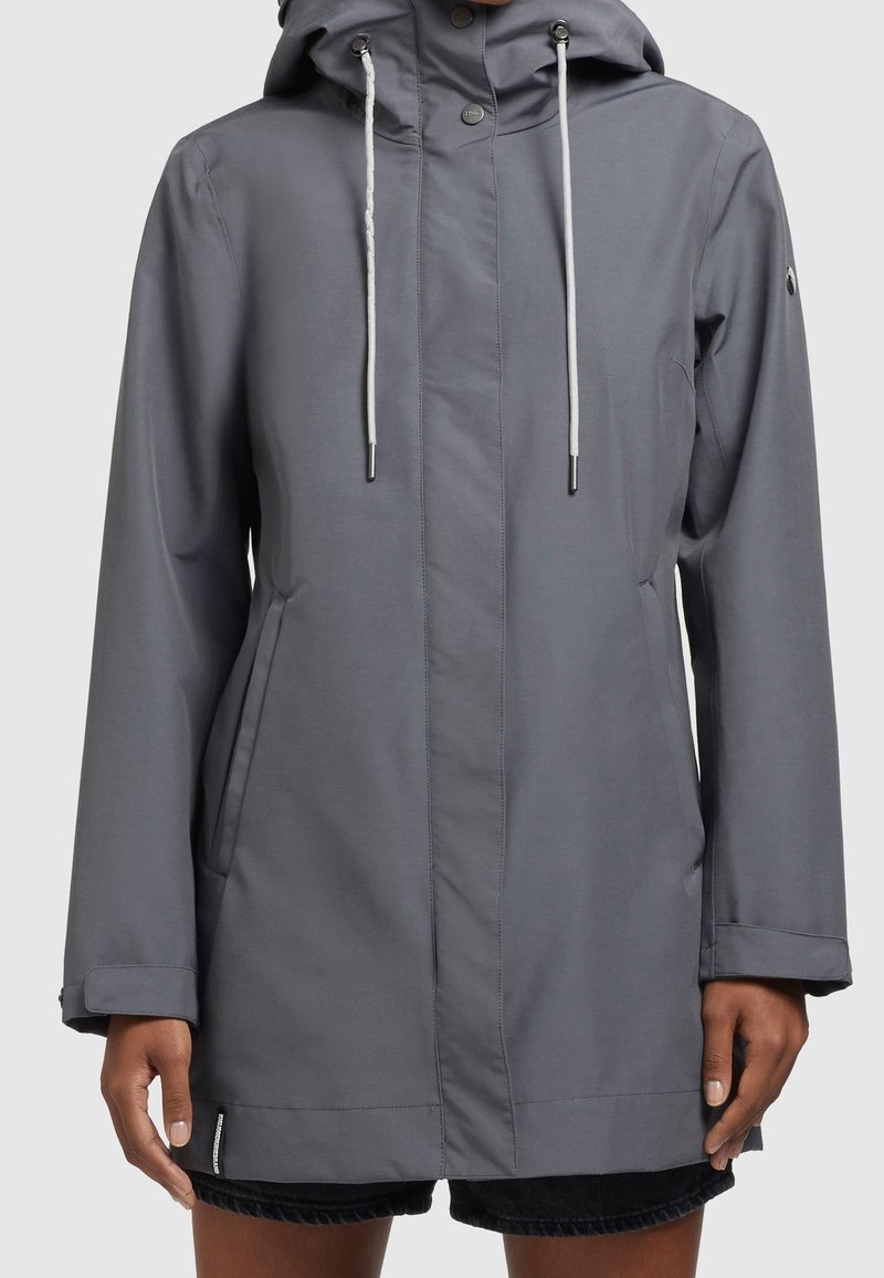 Šedá kapucňová bunda s bílými šňůrkami, předními kapsami a patentními knoflíky, nošená přes černé kraťasy na osobě s tmavou pletí.