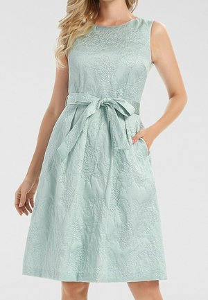 Robe sans manches bleu clair à motifs floraux texturés avec une ceinture nouée à la taille et des poches, portée par une femme debout aux cheveux blonds ondulés.