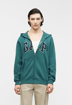 Ung mann med mørkt hår iført teal GAP hettejakke med glidelås over hvit skjorte og beige cargobukser, stående mot en ensfarget lys bakgrunn.