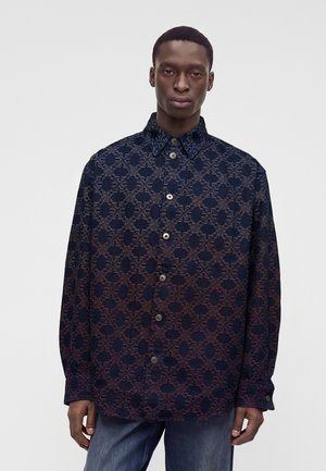 Homme portant une chemise sombre à boutons avec un motif géométrique complexe et un jean bleu, debout devant un fond clair uni.
