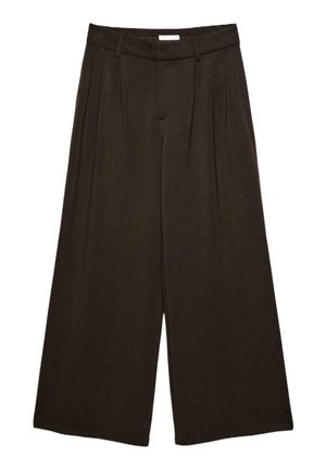 Femme portant un top court ample marron foncé et un pantalon à jambes larges, debout sur un sol en bois contre un mur blanc uni.