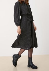 Žena oblečená v černých midi šatech s dlouhým rukávem a puntíky, s vypasovaným pasem a černými kotníkovými botami, kráčející po neutrální podlaze.