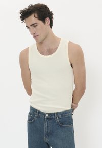 Giovane uomo con capelli ricci e scuri, che indossa una canottiera crema senza maniche e jeans blu, in piedi con le mani dietro la schiena su uno sfondo bianco.