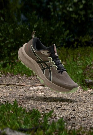 Chaussure de course marron et noire à semelle épaisse en l'air au-dessus d'un sentier rocheux entouré de végétation verte dans un environnement forestier.
