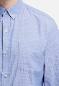 Vaaleansininen napillinen kauluspaita, jossa on hienovarainen pilkkukuosi, rintatasku ja valkoiset napit. Sileä kangas ja klassinen kaulus.