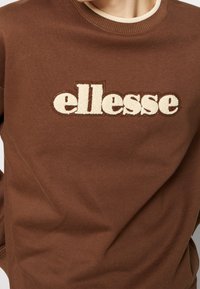 Hnědá mikina s krémovým žebrovaným výstřihem a vyšitým logem "ellesse" na hrudi. Měkká textura, volný střih, minimalistický design.