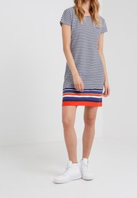 Gestreiftes Kleid in Marineblau und Weiß mit kurzen Ärmeln, das einen bunten gestreiften Saum in Rot, Blau und Gelb aufweist. Kombiniert mit weißen Sneakern.