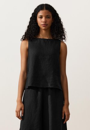 Kvinde med langt, krøllet hår iført en sort, ærmeløs hør-top og matchende løse sorte bukser, stående mod en simpel lys baggrund.