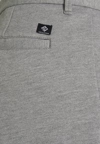 Pantaloni in tessuto grigio con motivo a spina di pesce, una tasca posteriore e un'etichetta nera con un logo. La texture appare liscia e tessuta.