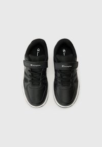 Chaussures de sport noires avec tige synthétique perforée, semelle intermédiaire blanche, lacets noirs et sangle Velcro réglable ; logo sur la sangle et sur la languette.