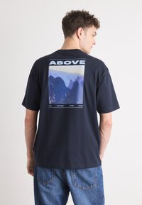 Tricou din bumbac bleumarin, cu un print grafic cu munți și textul "ABOVE" pe spate. Mâneci scurte, croială lejeră și guler rotund.