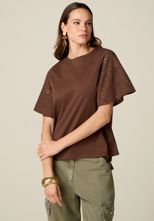 Donna con top marrone con maniche in pizzo traforato, pantaloni cargo verde oliva, grande bracciale d'oro e orecchini pendenti su sfondo semplice.