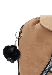 Mochila de nylon color marrón con un bolsillo frontal con cremallera, cierre con solapa y un llavero de peluche negro en forma de mono.