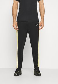 Schwarze Sporthosen mit gelben seitlichen Streifen, elastischem Taillenbund und Logo am Hüftbereich, aus einem glatten, dehnbaren Material gefertigt.