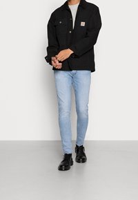 Persona vistiendo una chaqueta negra con botones, jeans ajustados de color azul claro, zapatos de cuero negros, de pie sobre un suelo gris contra una pared blanca.