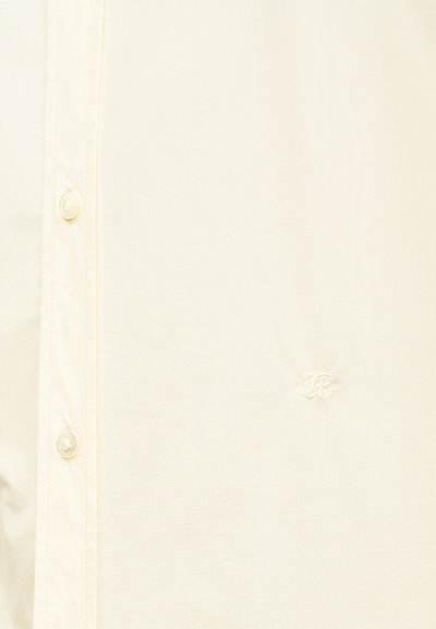 Koszula zapinana na guziki w kolorze kremowym o gładkiej fakturze, z okrągłymi guzikami i małym haftowanym logo w pobliżu środka.