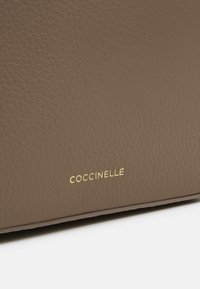 Sac à main en cuir marron avec une finition texturée, arborant un logo "COCCINELLE" en relief doré sur une surface lisse.