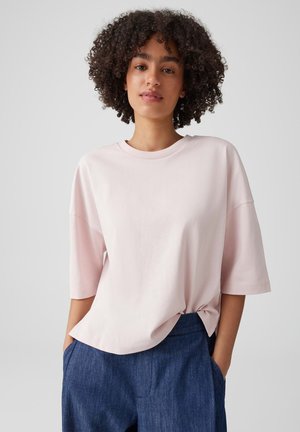 Junge Person mit lockigem Haar, die ein hellrosa oversized T-Shirt und blaue Hosen trägt, steht mit den Händen in den Taschen vor einem einfarbigen Hintergrund.
