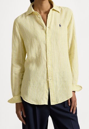 Chemise jaune à manches longues avec de fines rayures verticales blanches et bleues, portée avec un pantalon sombre, montrant le logo sur la poitrine gauche.