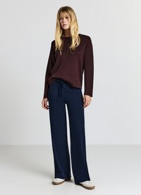 Haut marron à manches longues accompagné d'un pantalon large bleu marine, avec une taille à cordon. Le modèle porte des chaussures blanches avec une semelle marron clair.