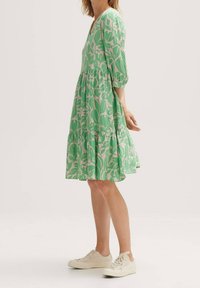 Kobieta w zielonej i białej kwiecistej sukience do kolan z rękawami 3/4 i białych sneakersach, stojąca bokiem na jednolitym tle.