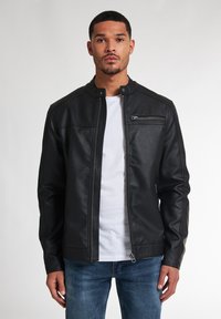 Zwarte nepleer jacket met een hoge kraag, voorrits en een ritszak op de borst. Gekozen boven een wit T-shirt en blauwe jeans.
