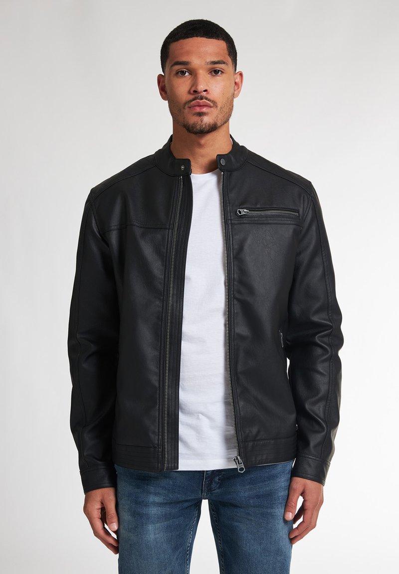 Zwarte nepleer jacket met een hoge kraag, voorrits en een ritszak op de borst. Gekozen boven een wit T-shirt en blauwe jeans.