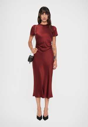 Vestido de seda bordeaux com mangas curtas, detalhe franzido na cintura, saia fluida, combinado com sapatos de salto agudo pretos e uma pequena bolsa de ombro.